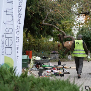 Widok ubranego roboczo mężczyzny od tyłu, który niesie skrzynkę  obok ułożonych na podjeździe ogrodowym licznych narzędzi ogrodowych.