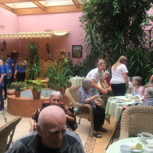 Seniorzy przy ogrodowych stolikach delektujący się kawą, a pośród nich wolontariusze