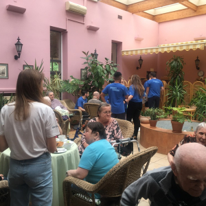 Seniorzy przy ogrodowych stolikach delektujący się kawą, a pośród nich wolontariusze