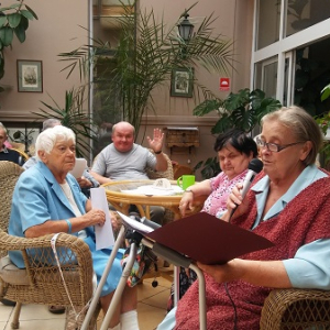 Seniorzy siedzący w kawiarence przy stolikach oraz recytująca poezję kobieta