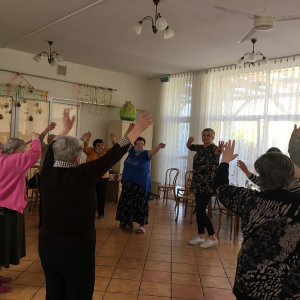 Grupa Seniorów stojąca w okręgu podczas zajęć ruchowych z uniesionymi rękoma.