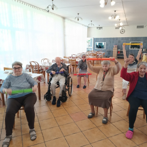 Seniorzy siedzący na krzesłach w czasie ćwiczeń z makaronami z gąbki.